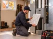 An HVAC technician kneels, holding a clean furnace filter, preparing to install it into a grey furnace unit, a key step in fall furnace maintenance.
